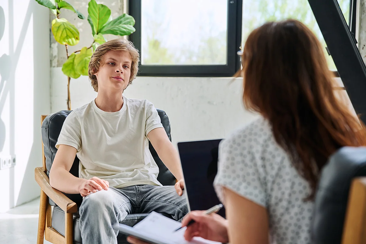 A teenage boy sits across from a therapist in a bright office, appearing relaxed and engaged in conversation.