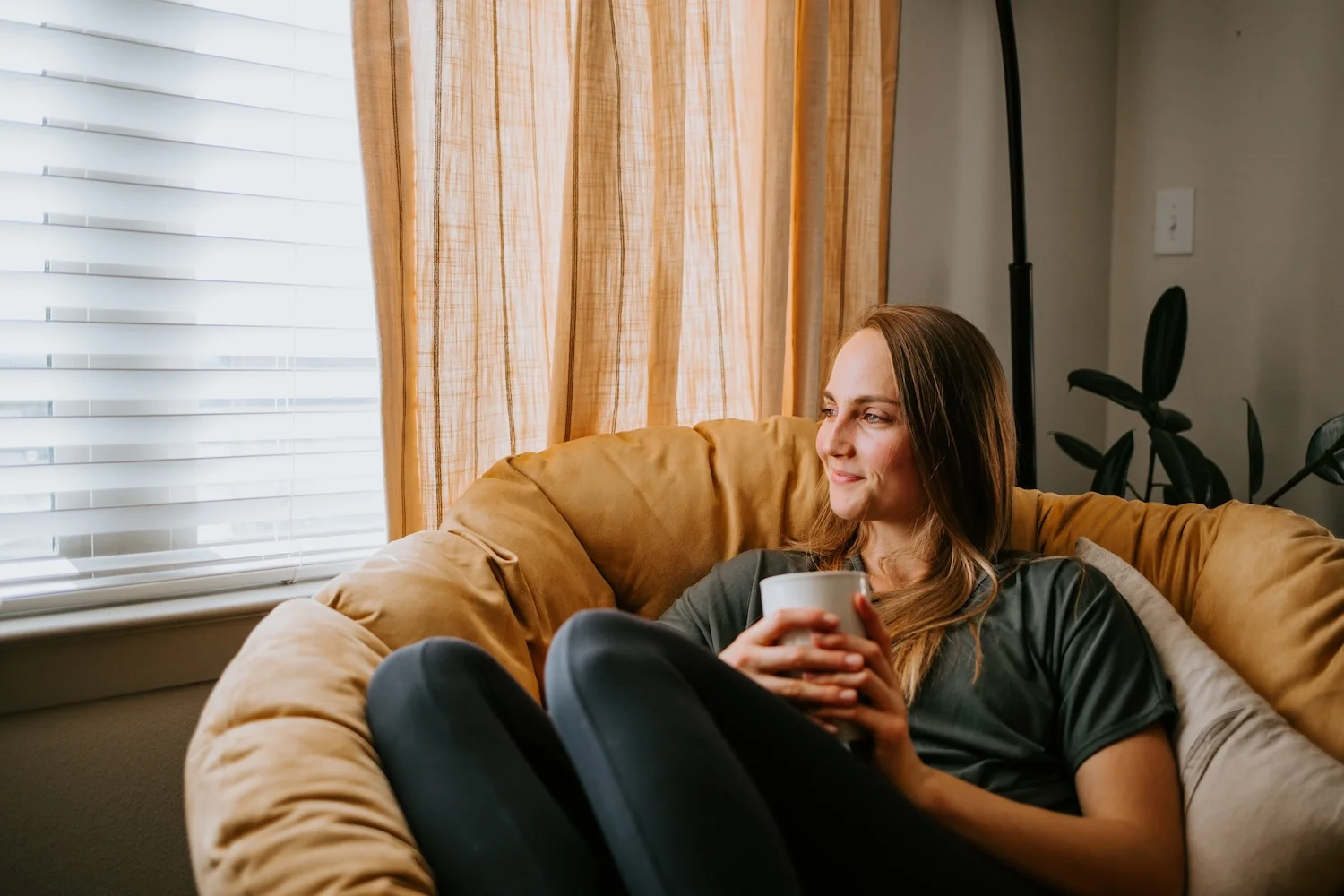 A woman sits curled up in a soft tan armchair, holding a mug with both hands and looking peacefully out a sunlit window. Warm curtains glow behind her, creating a cozy, relaxed atmosphere.