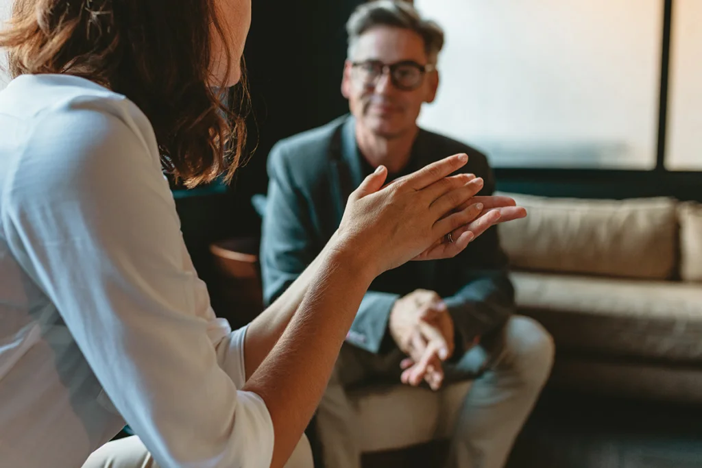 A person gestures while speaking to a therapist seated nearby in a warmly lit office.