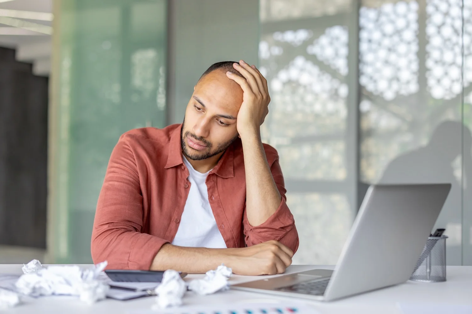 Man sitting at a desk with a laptop, looking stressed and resting his head on his hand, surrounded by crumpled papers, suggesting frustration or mental fatigue at work.