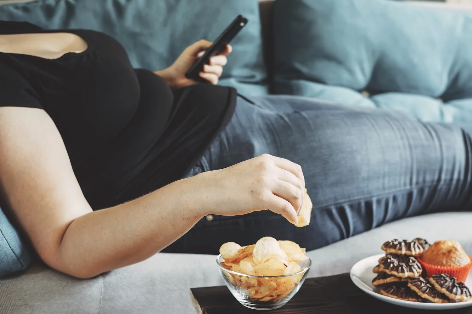 Person lying on a couch holding a TV remote and reaching for a bowl of potato chips, with a plate of pastries nearby, suggesting relaxation or boredom eating while watching TV.