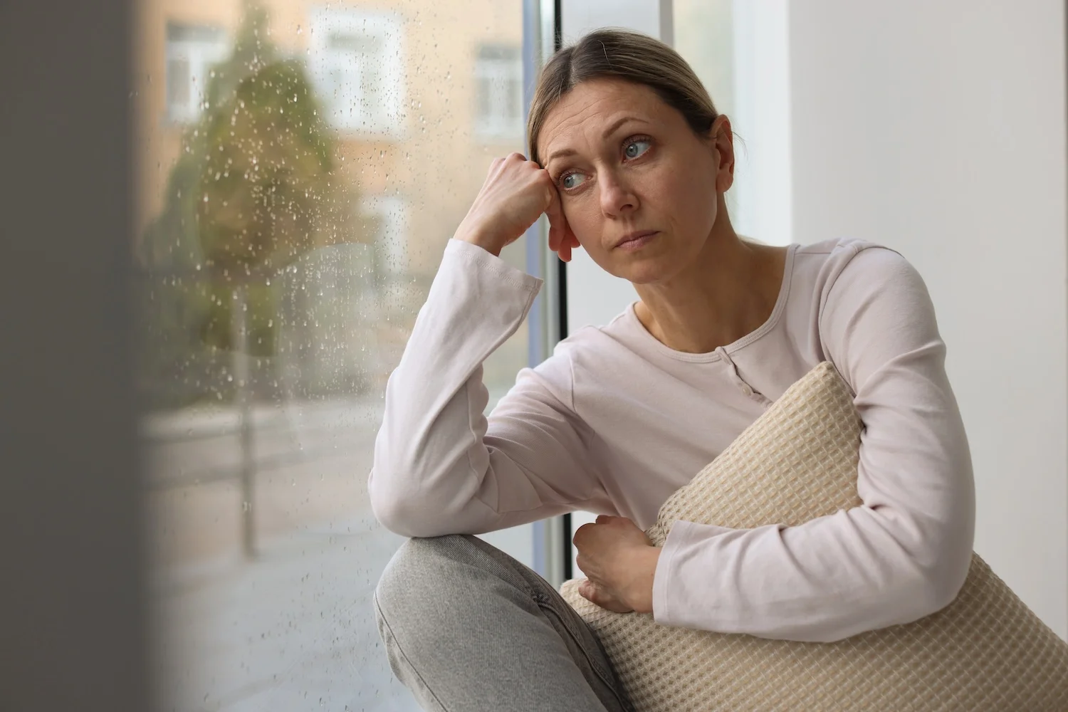 A woman sits by a window on a rainy day, resting her head on her hand and holding a pillow, appearing thoughtful or concerned.