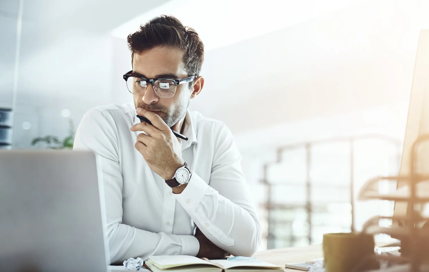 Man wearing glasses and a white shirt sitting at a desk, thoughtfully looking at a laptop screen with a pen near his mouth, suggesting focus or contemplation while working.