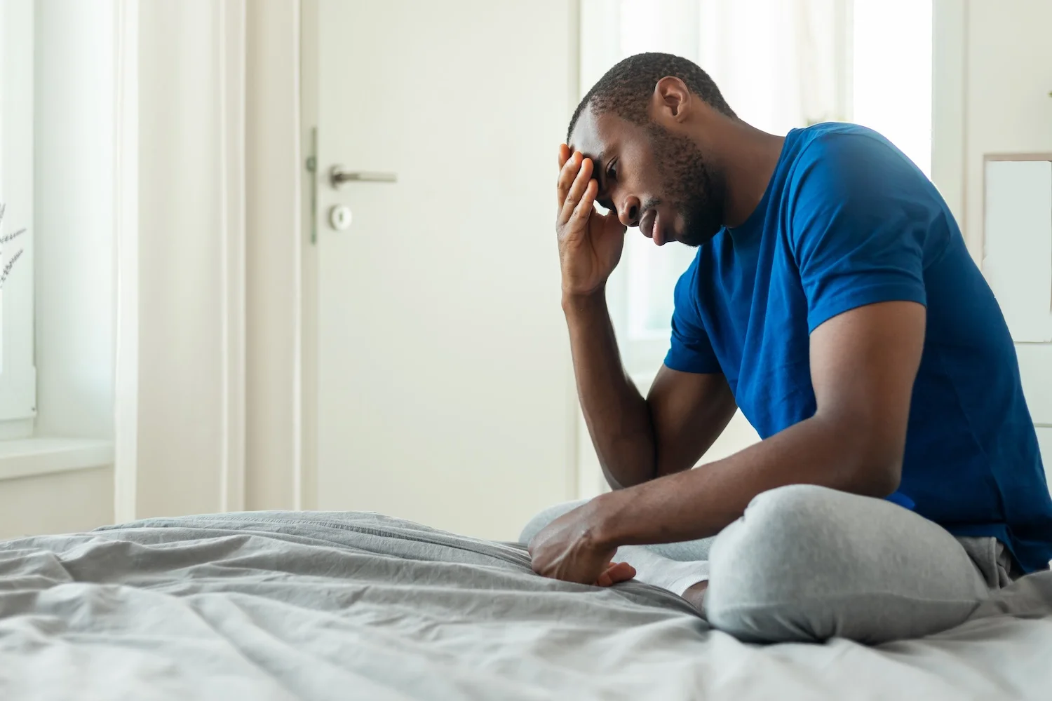 A man in a blue shirt sits on the edge of his bed with his hand on his forehead, showing signs of stress or exhaustion.