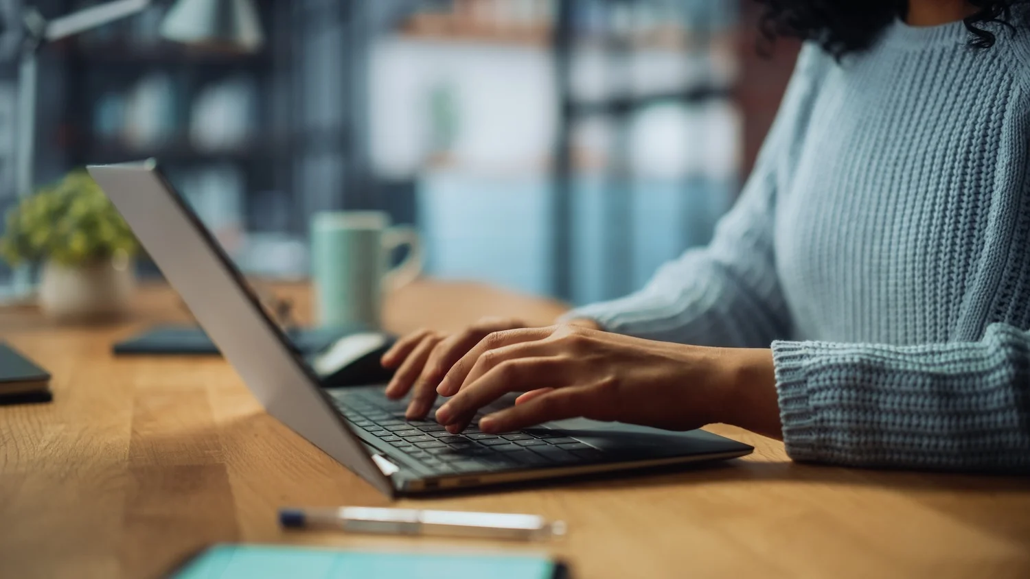 Close-up of a person in a light blue sweater typing on a laptop at a wooden desk, with a blurred background of a cozy home office.
