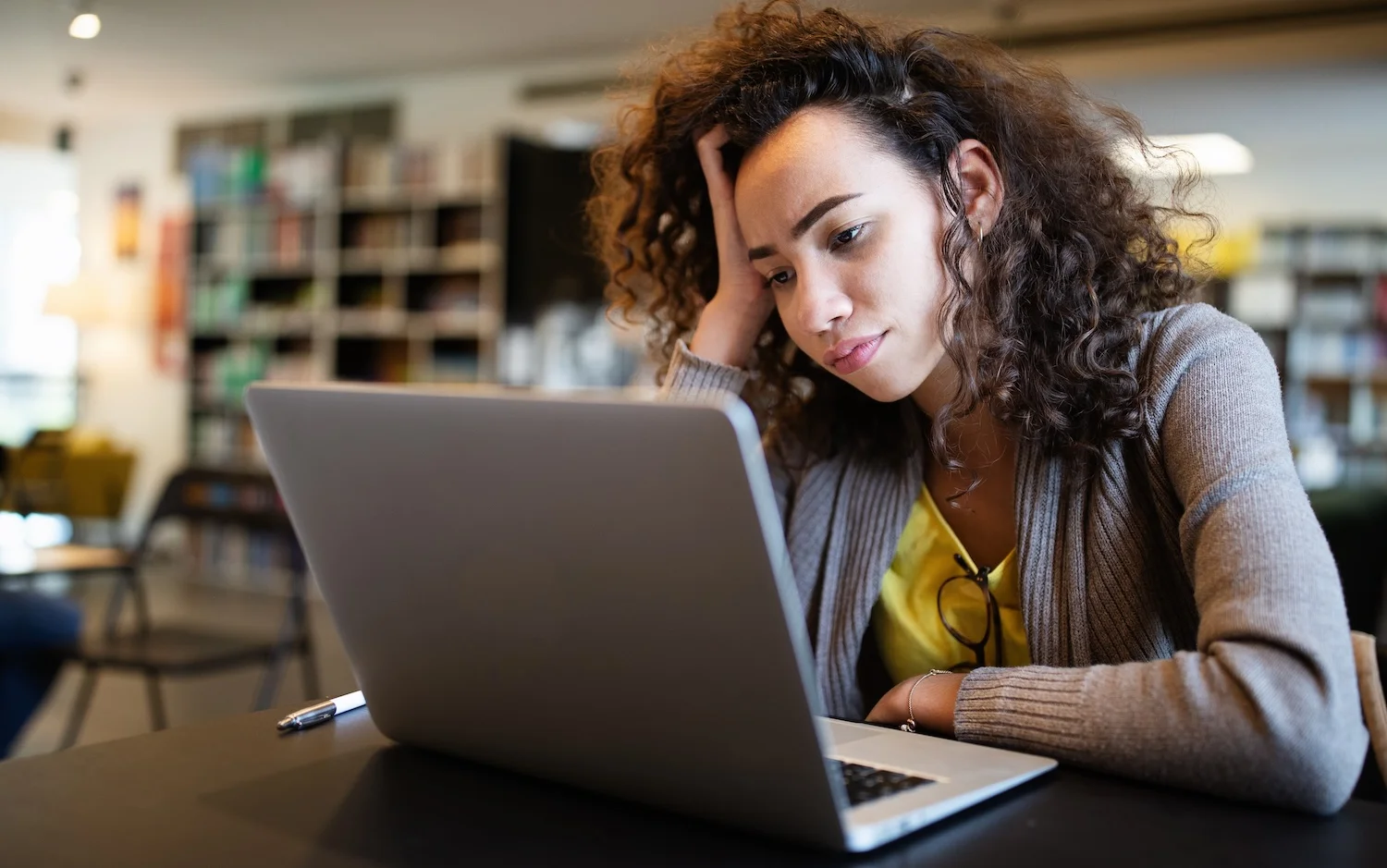 Woman sitting at a table with a laptop, resting her head on her hand and looking at the screen with a thoughtful or tired expression, suggesting concentration or frustration while working or studying.