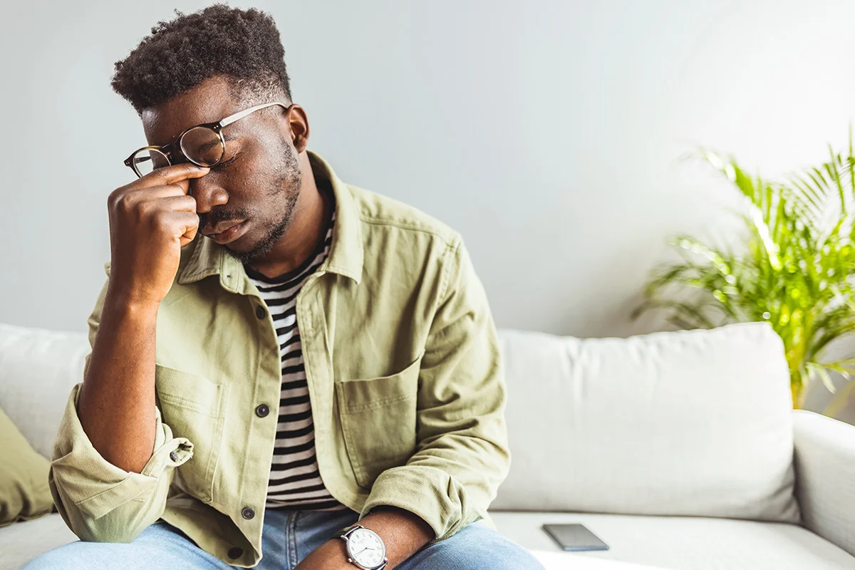 A man in glasses sits on a couch with his eyes closed and hand to his forehead, showing fatigue or frustration.