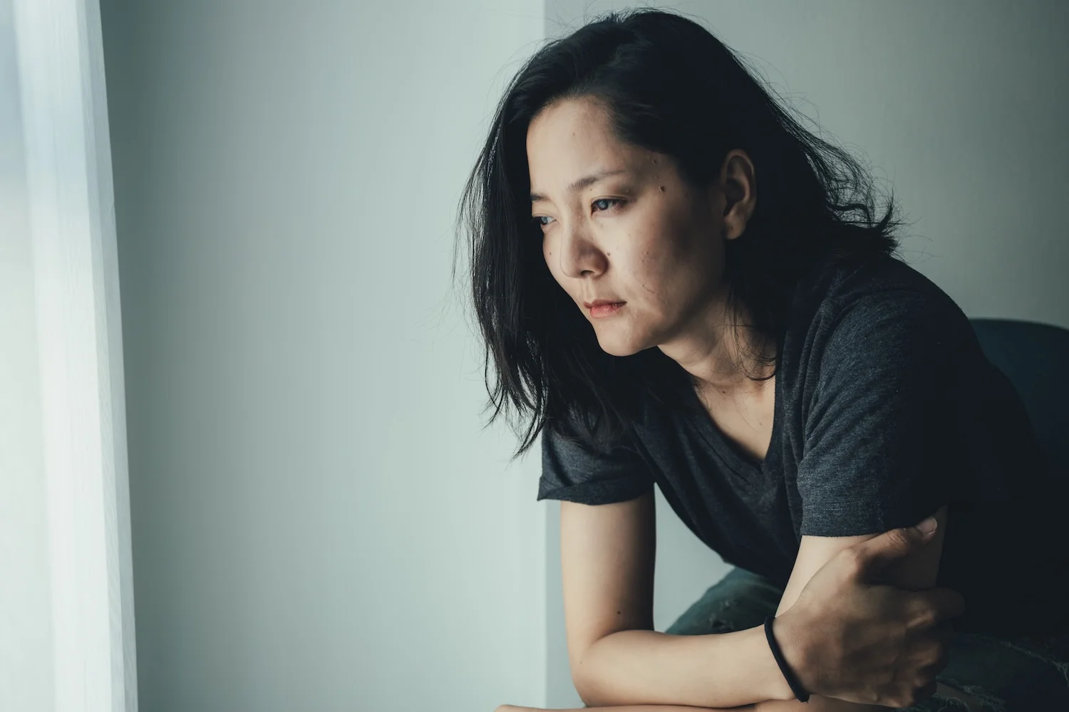 A woman sitting indoors with a somber expression, leaning forward and looking out a window, conveying feelings of sadness or deep thought.
