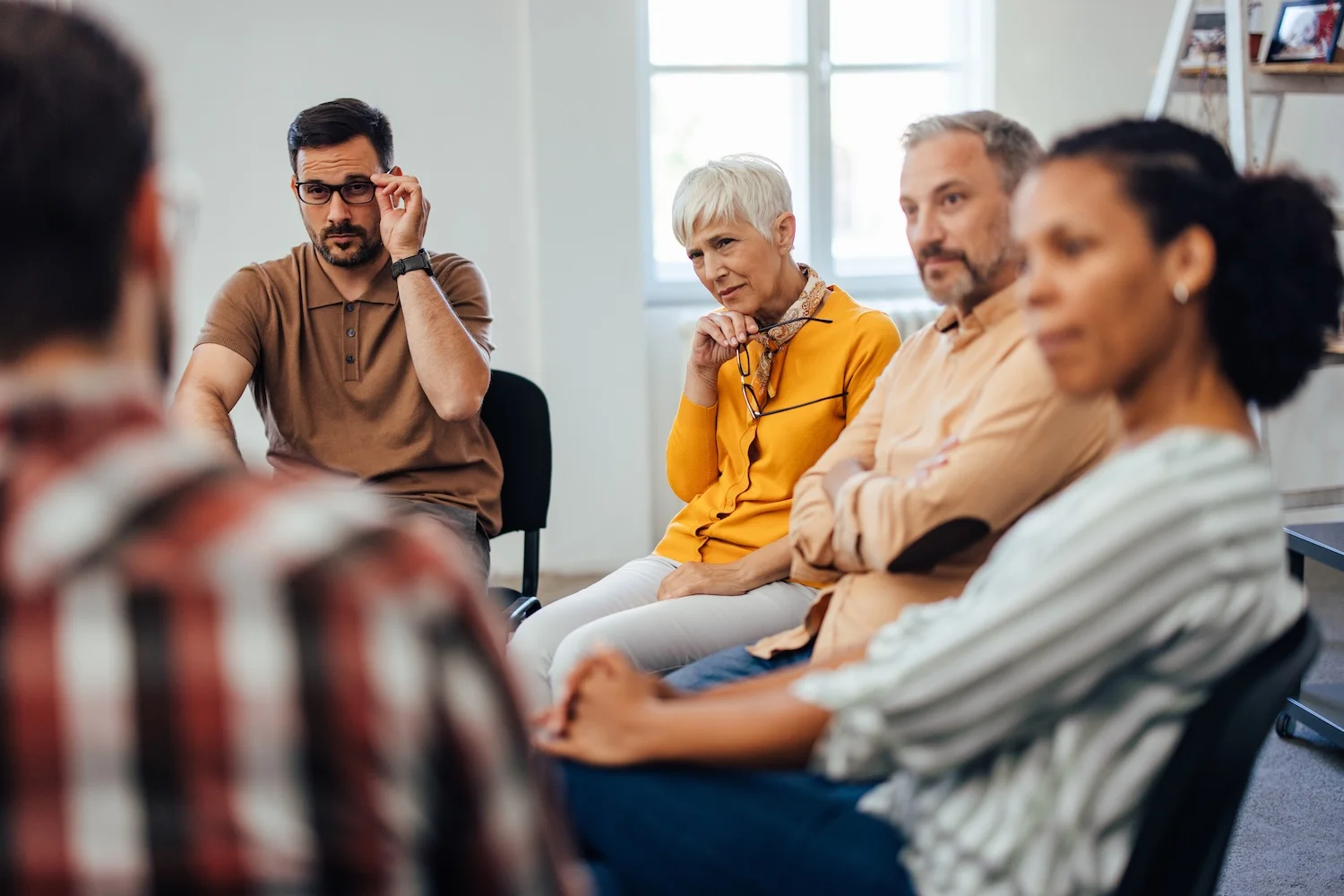 A small group of adults sits in a circle during what appears to be a support or discussion meeting, listening attentively to someone speaking off-camera. The group includes men and women of different ages, all seated on chairs in a bright room with a window in the background.