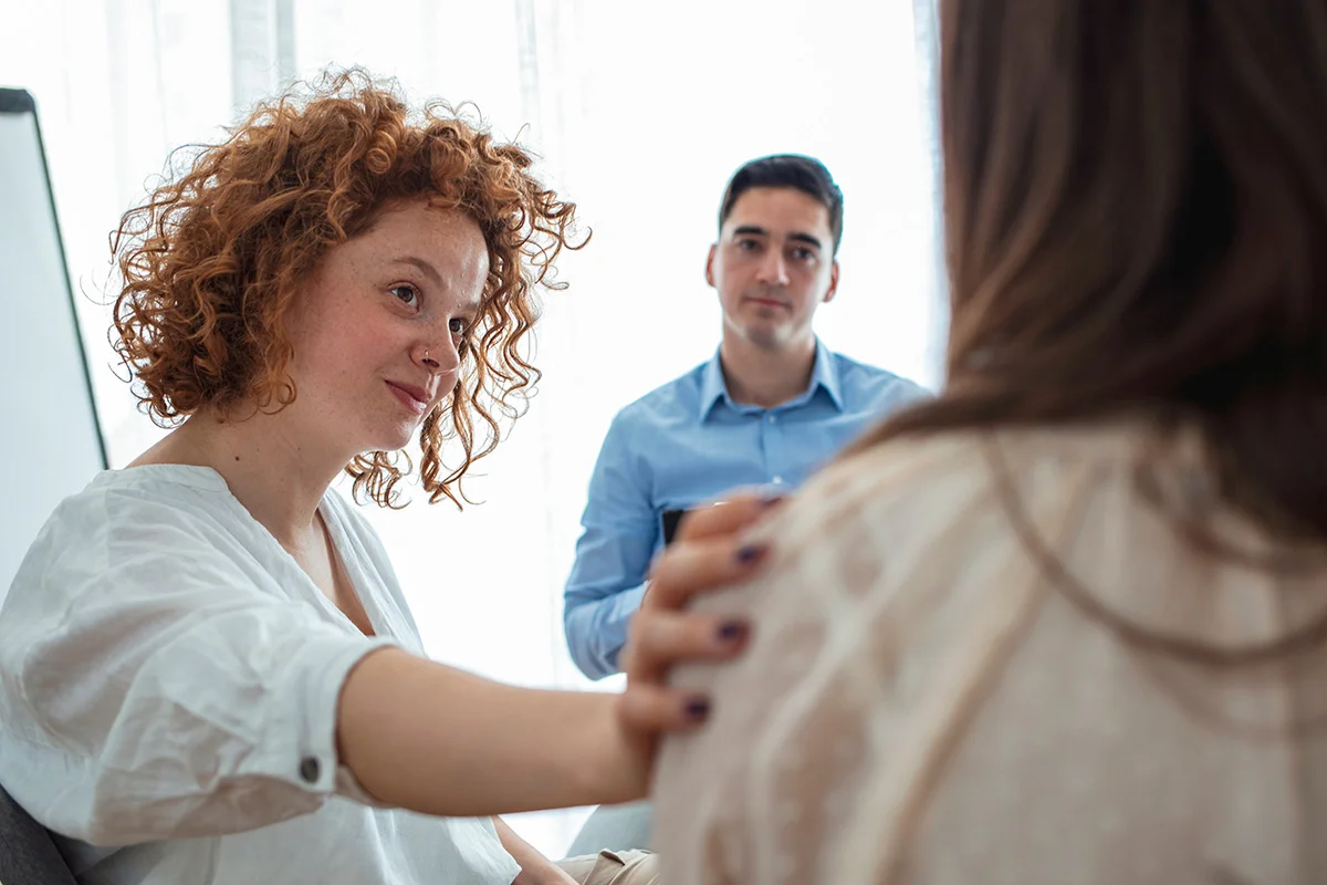 A woman reaches out to comfort another person in a small group session, with a man observing supportively.